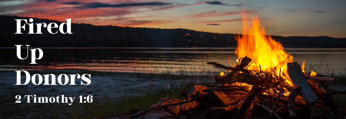 Campfire burning brightly by a lake at sunset with the words “Fired Up Donors” and “2 Timothy 1:6” written on the left side.