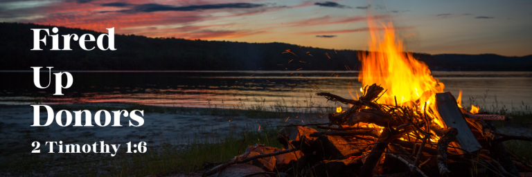 Campfire burning brightly by a lake at sunset with the words “Fired Up Donors” and “2 Timothy 1:6” written on the left side.