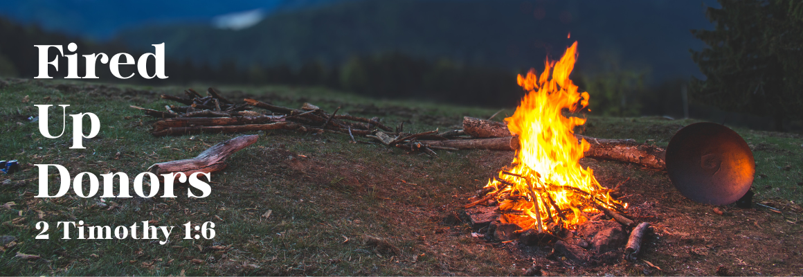 Campfire burning brightly in an open grassy field with mountains in the background, next to text that reads 'Fired Up Donors – 2 Timothy 1:6'.
