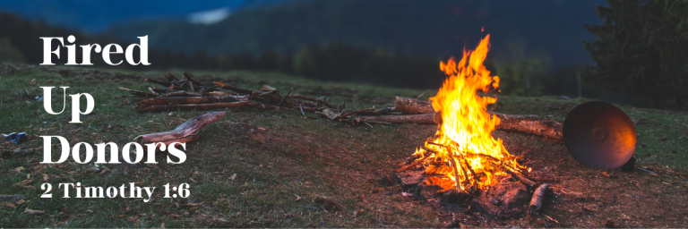 Campfire burning brightly in an open grassy field with mountains in the background, next to text that reads 'Fired Up Donors – 2 Timothy 1:6'.