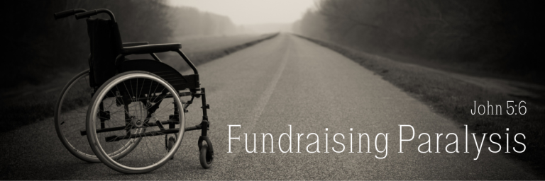 An empty wheelchair sitting on a long empty road disappearing into a foggy horizon, in black and white, with the text Fundraising Paralysis and the scripture reference John 5:6 in white lettering.