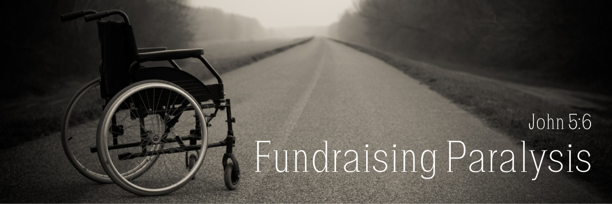 An empty wheelchair sitting on a long empty road disappearing into a foggy horizon, in black and white, with the text Fundraising Paralysis and the scripture reference John 5:6 in white lettering.